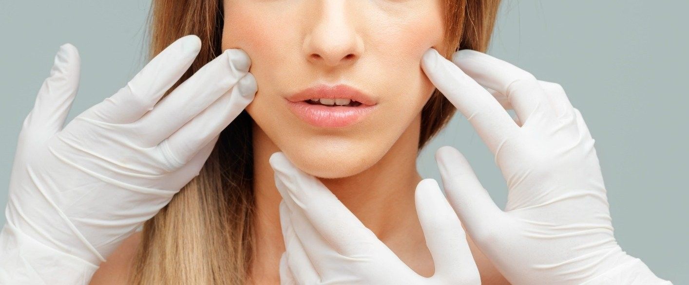 Manos enguantadas examinando el rostro de una mujer durante un tratamiento cosmético.