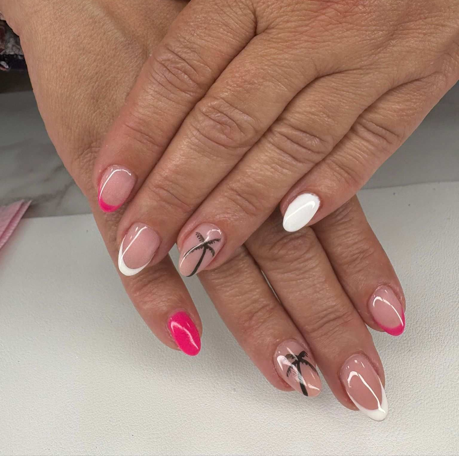 Manos con diseños de uñas en rosa, blanco y color nude, con motivos de cintas negras, sobre una mesa de luz.