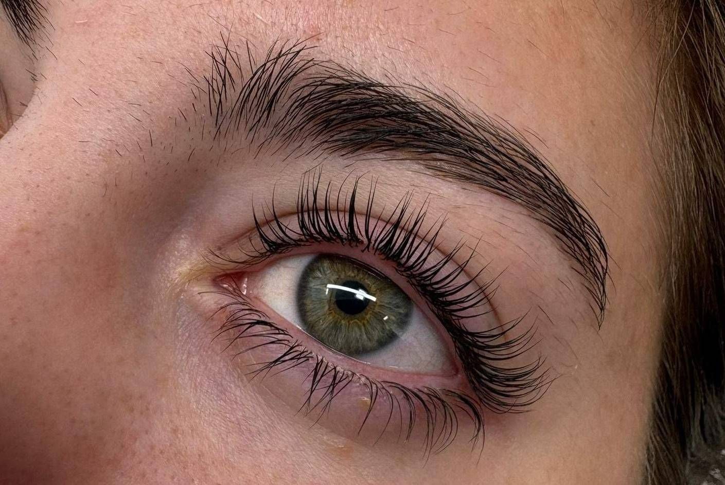 Primer plano de un ojo marrón verdoso con pestañas largas y ceja oscura bajo luz natural.