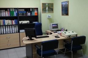 Escritorio de oficina con ordenador, estanterías con carpetas y dos sillas en un pequeño espacio de trabajo.