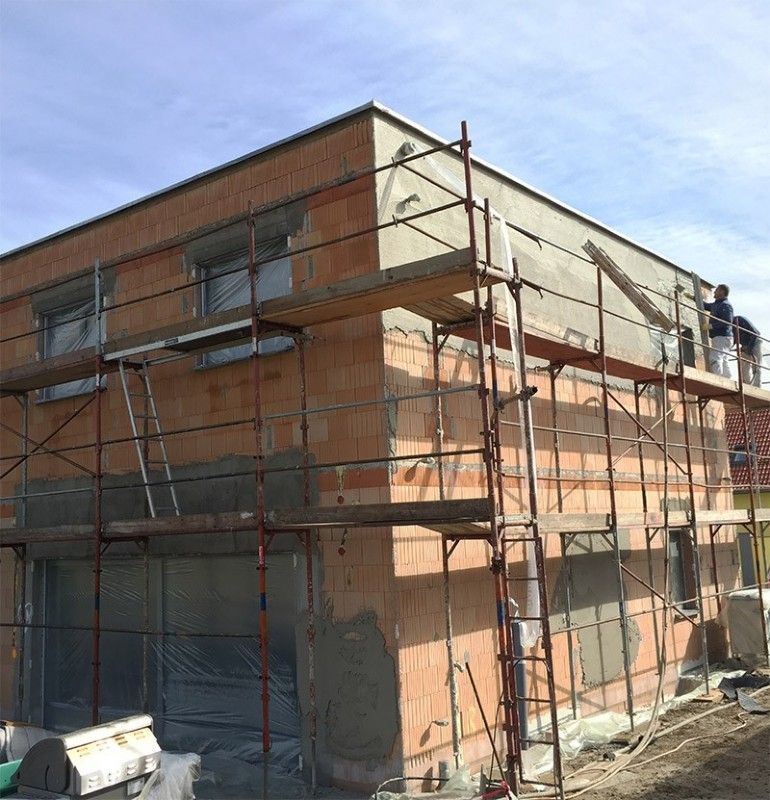 Ein zweistöckiges Backsteingebäude im Bau, umhüllt von einem Metallgerüst, während ein Arbeiter die Außenfassade verputzt.