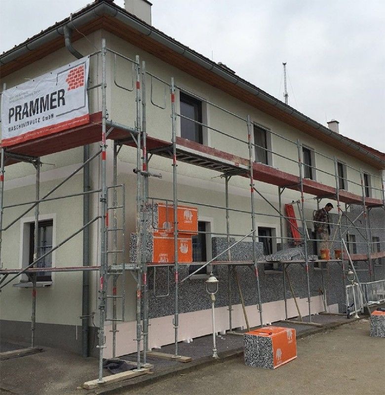 Ein Gerüst umgibt ein Gebäude, das derzeit renoviert wird; Arbeiter montieren graue Dämmplatten an der unteren Außenfassade.