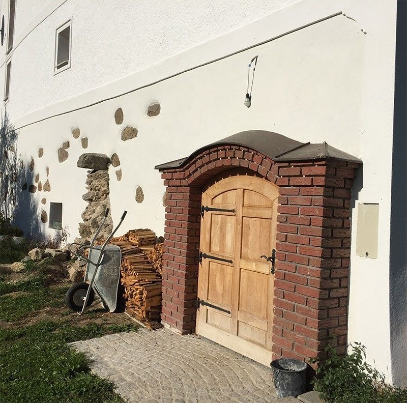 Ein roter Backsteinbogen mit hölzernen Doppeltüren, eingefasst in eine weiße Mauer mit verstreuten Steinverzierungen und Brennholz.