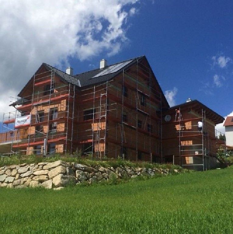 Ein mehrstöckiges Haus im Bau, umgeben von Gerüsten, vor der Kulisse eines grünen Hügels unter blauem Himmel.