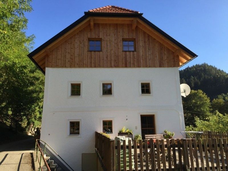 Ein weißes, dreistöckiges Haus mit hölzernem Dachgiebel, braunem Ziegeldach und umzäunter Holzterrasse in waldreicher Umgebung.