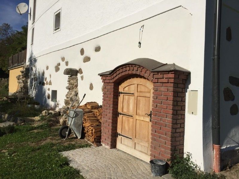 Ein gewölbter, roter Backsteineingang führt in eine weiße Gebäudewand mit Ziersteinen, neben der ein Holzstapel und eine Schubkarre stehen.