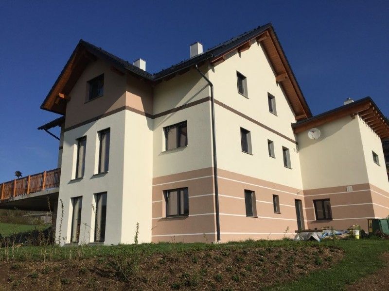 Ein zweistöckiges Haus mit beigen und pfirsichfarbenen Wänden, dunkel gerahmten Fenstern und einem braunen Giebeldach unter einem klaren blauen Himmel.