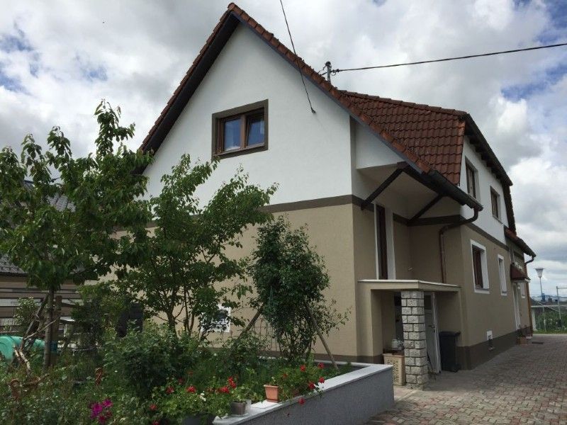 Ein zweistöckiges Haus mit weiß-beiger Fassade und braunem Ziegeldach, gesehen von einem seitlichen Gartenweg aus.
