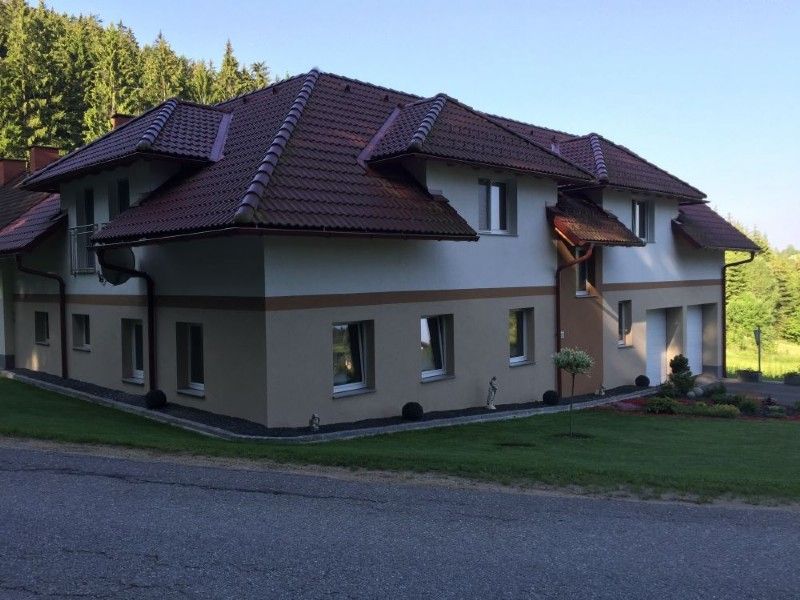 Ein zweistöckiges Haus mit braunem Ziegeldach, weißen und beigen Außenwänden und einer Auffahrt, vor dem Hintergrund einer von Bäumen gesäumten Kulisse.