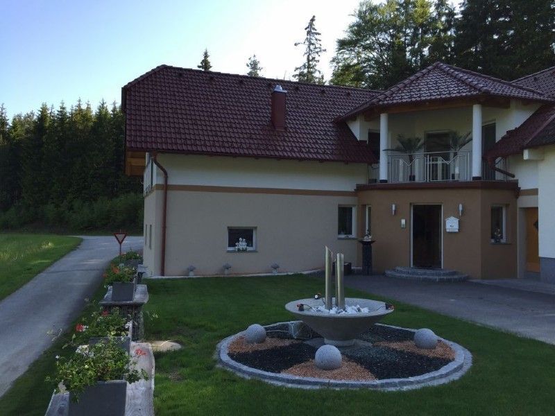 Ein zweistöckiges Gebäude mit braunem Ziegeldach, beigen Wänden und einem modernen, mehrstufigen Brunnen im Vorgarten.