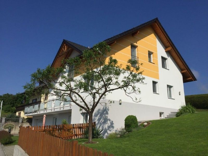 Ein zweistöckiges Haus mit weiß-gelben Wänden, einem dunklen Dach und einem Holzzaun, umgeben von einem grasbewachsenen Rasen unter blauem Himmel.