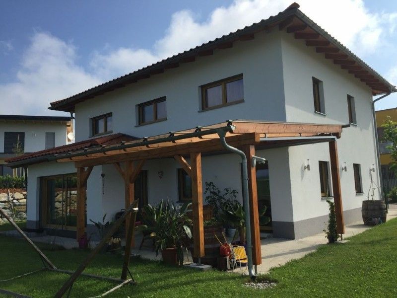 Ein weißes, zweistöckiges Haus mit einer überdachten Holzterrasse, umgeben von einem grünen Rasen an einem sonnigen Tag.