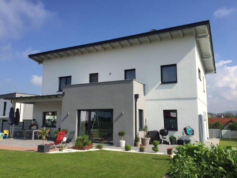 Ein zweistöckiges weißes Haus mit einem grauen Mittelteil, dunklen Fensterrahmen, einer Terrasse und einem grasbewachsenen Hinterhof unter blauem Himmel.