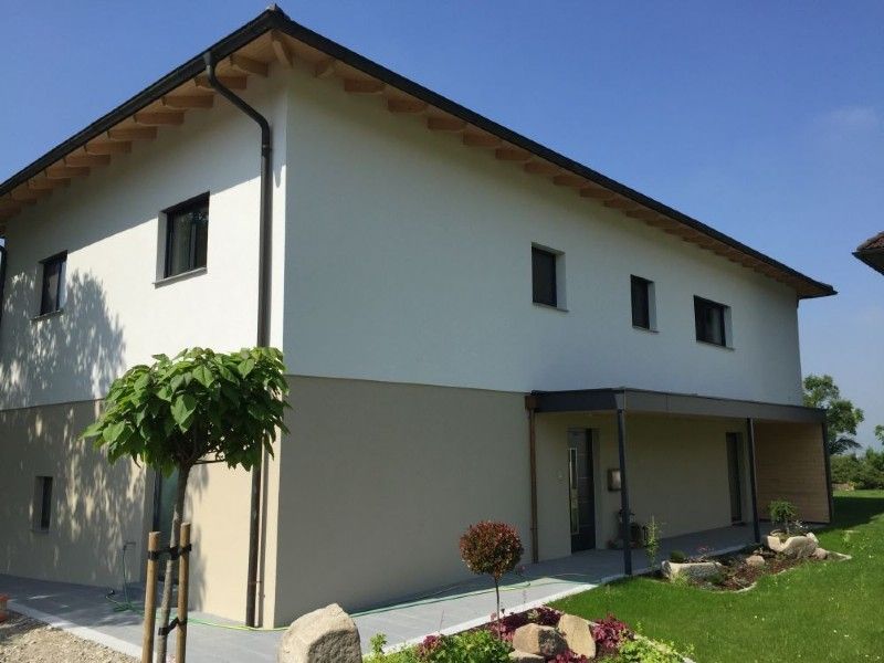 Ein zweistöckiges Haus mit weißem Obergeschoss und beigem Untergeschoss, mit überdachter Terrasse und einem kleinen, gepflegten Garten.