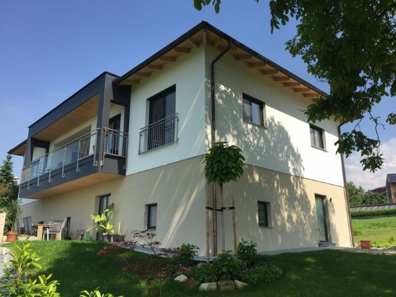 Ein zweistöckiges Haus mit weiß-beiger Fassade, einem Balkon und einem grünen Rasen unter klarem, blauem Himmel.