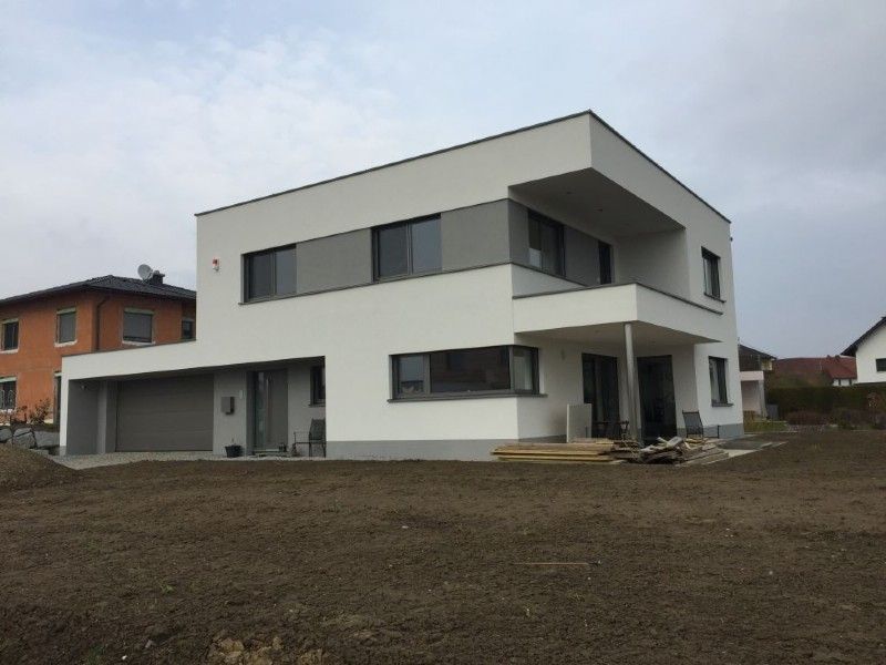 Ein modernes, zweistöckiges weißes Haus mit Flachdach, grauen Akzenten, einer Garage und einem unfertigen Garten unter bedecktem Himmel.