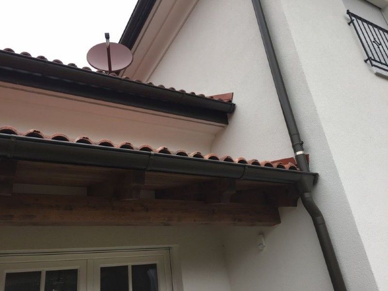 Eine Ansicht aus der Froschperspektive auf die Außenfassade eines weißen Gebäudes mit einem Ziegeldachüberstand, braunen Holzbalken und einem dunklen Fallrohr.