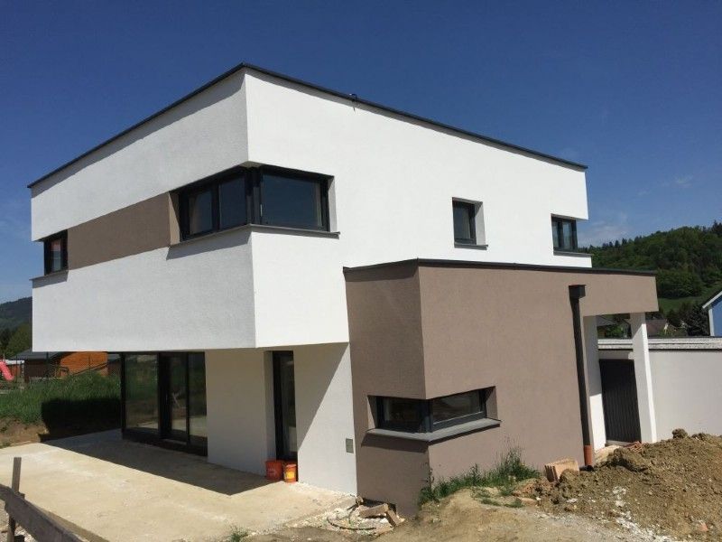 Ein modernes zweistöckiges Haus mit weiß-beigen Außenwänden, Flachdach und einer ebenerdigen Terrasse unter strahlend blauem Himmel.