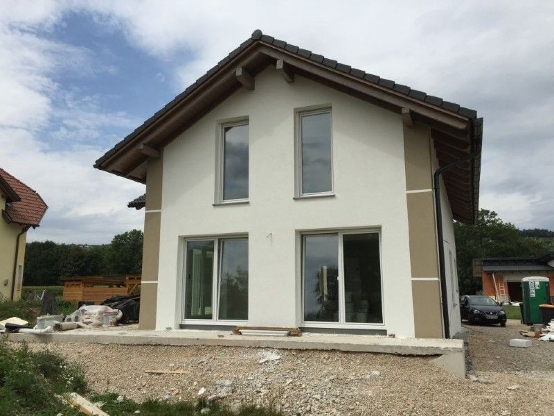 Ein neu gebautes zweistöckiges Haus mit weißen Wänden, hellbraunen Eckakzenten, einem braunen Dach und großen Fenstern, umgeben von Kies.