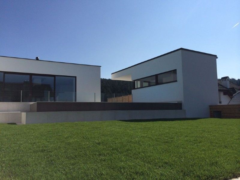 Ein modernes, minimalistisches weißes Haus mit großen Glasfenstern, einer dunklen Terrasse und einem gemähten Rasen unter klarem, blauem Himmel.