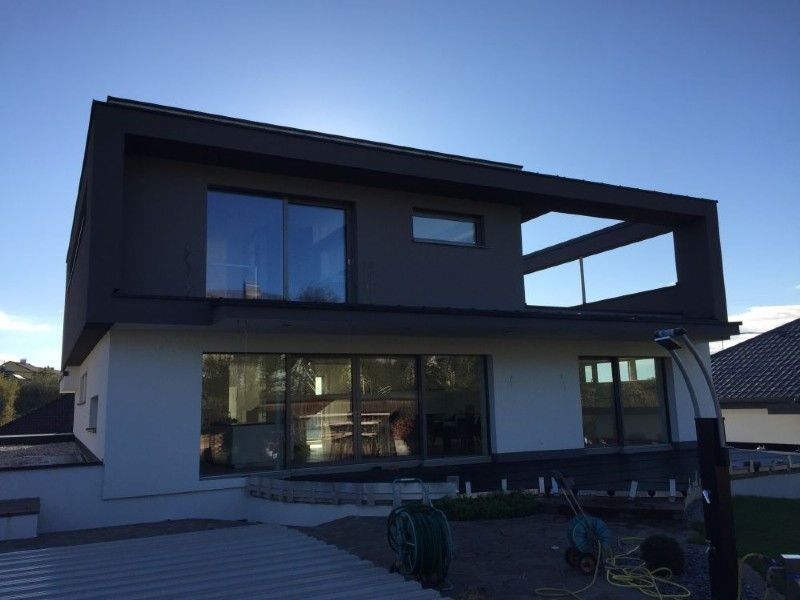 Ein modernes zweistöckiges Haus mit weißem Untergeschoss, dunkelgrauem Obergeschoss, großen Glasfenstern und einem offenen Balkon.