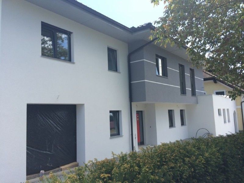 Ein modernes zweistöckiges Haus mit weißer Fassade, dunkelgrauem Obergeschoss, schwarzem Garagentor und grüner Hecke im Vorgarten.