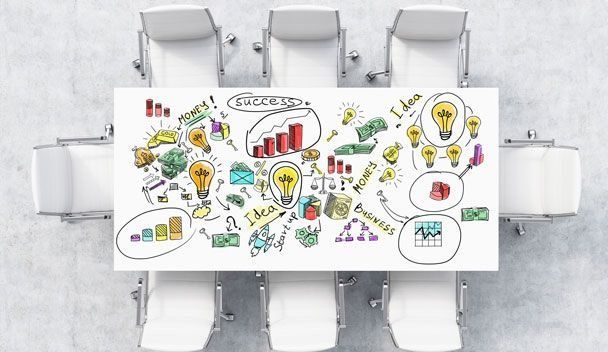 Vista aérea de una mesa de reuniones con sillas blancas. La mesa tiene un plan de negocios dibujado con diagramas y bombillas.