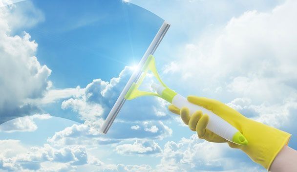 Una mano en un guante amarillo utiliza una escobilla de goma para limpiar una ventana, con cielo azul y nubes en el fondo.