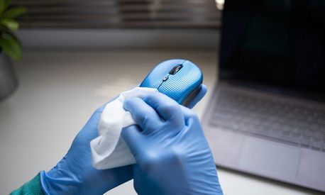 Una persona que lleva guantes azules limpia un ratón de ordenador azul con una toallita blanca, cerca de una computadora portátil.