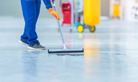 Persona trapeando un piso brillante con guantes naranjas; carrito de productos de limpieza al fondo.