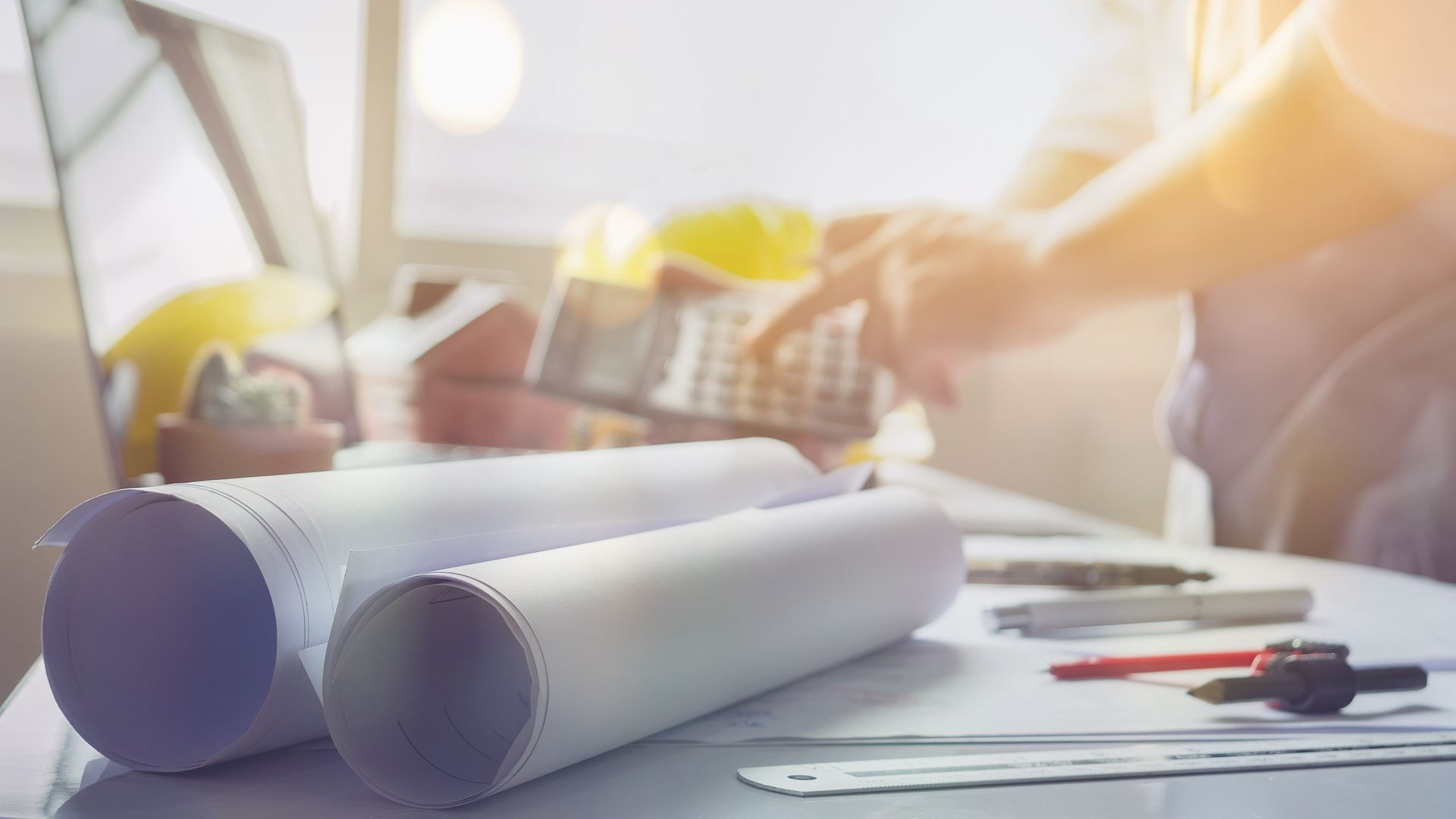 Des plans et une calculatrice sont posés sur un bureau, avec une personne floue en train de calculer en arrière-plan.