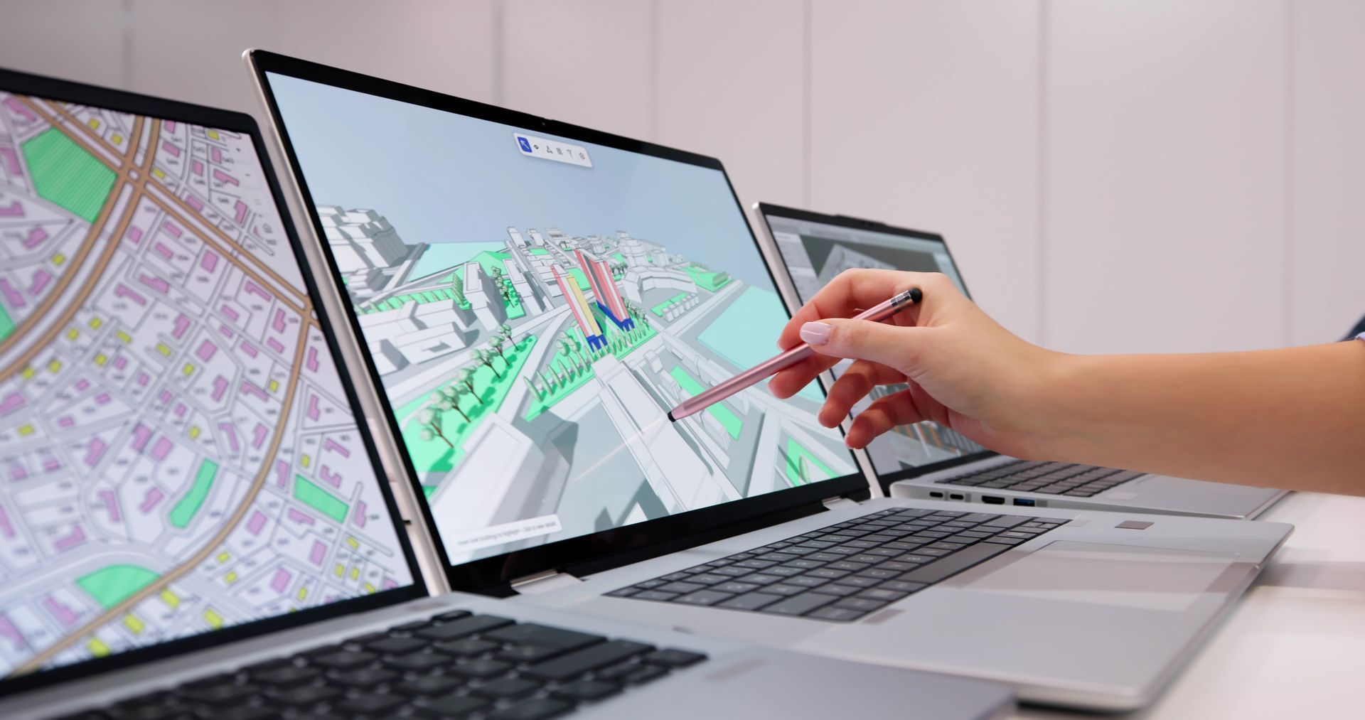 Trois ordinateurs portables affichent des cartes. Une main munie d'un stylet interagit avec l'écran central.