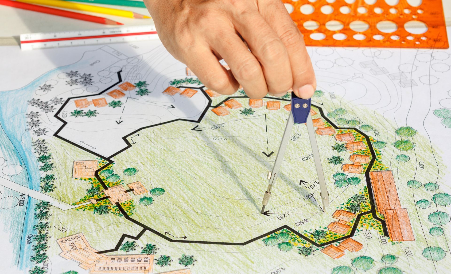 Main utilisant un compas sur un plan d'architecture paysagère coloré représentant des maisons et des arbres.