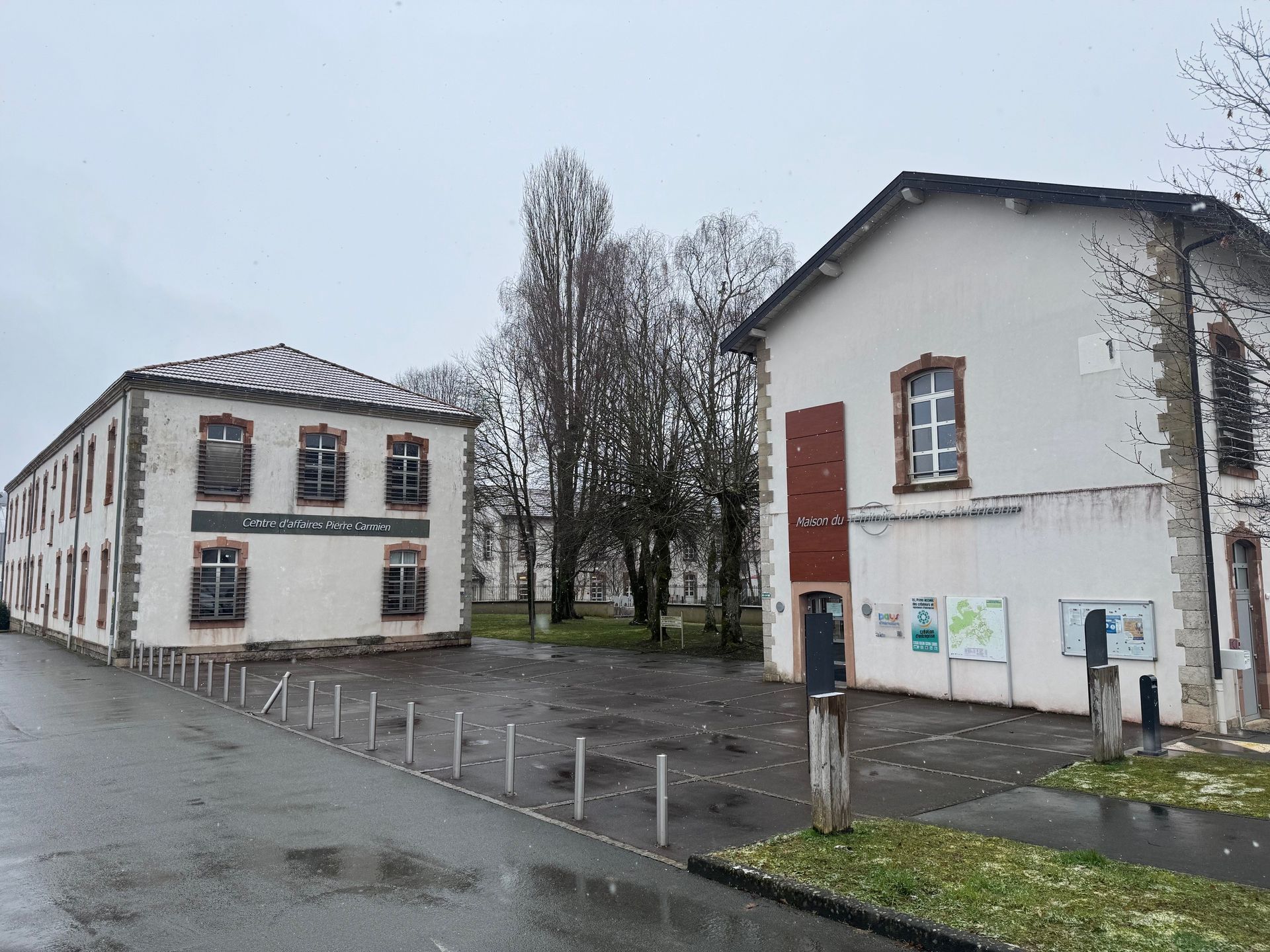 Deux bâtiments clairs aux boiseries sombres par temps nuageux. Une route et des arbres dénudés les séparent.