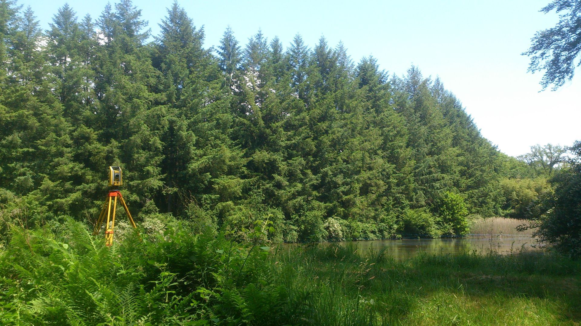 Un instrument de topographie installé près d'un plan d'eau, devant une dense rangée d'arbres.