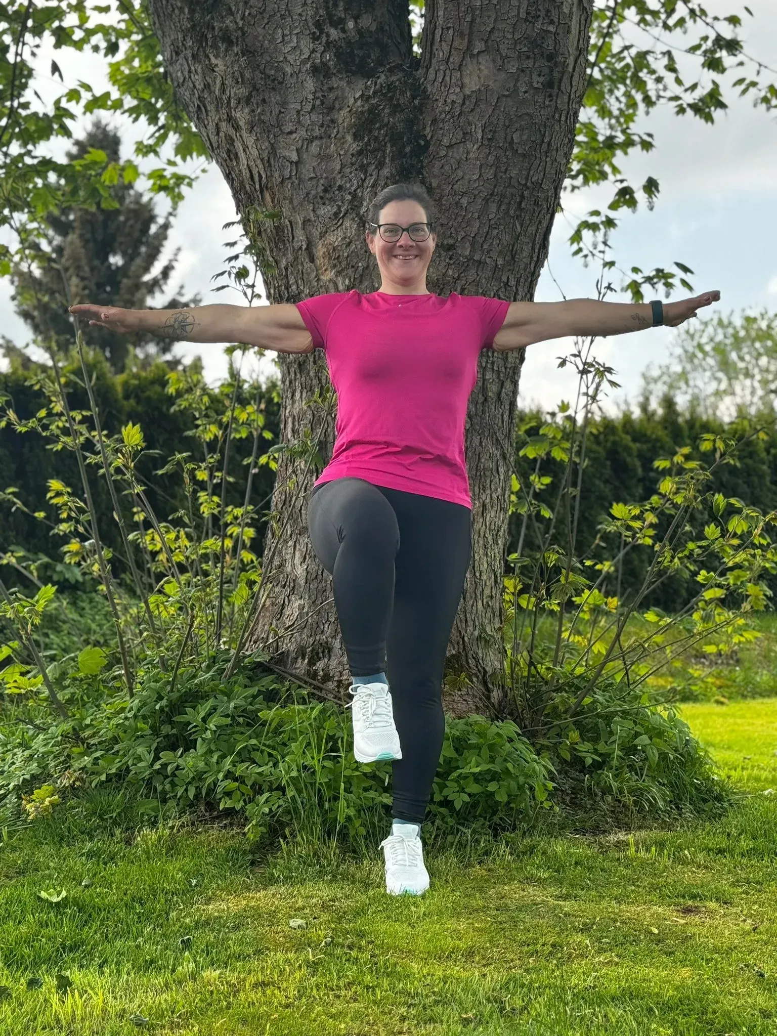 Eine Person balanciert auf einem Bein vor einem Baum, die Arme zur Seite ausgestreckt.