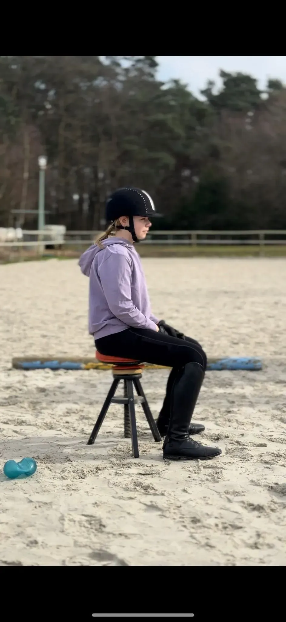 Eine Person mit Helm und lila Kapuzenpullover sitzt auf einem Hocker in einer Reithalle im Freien.