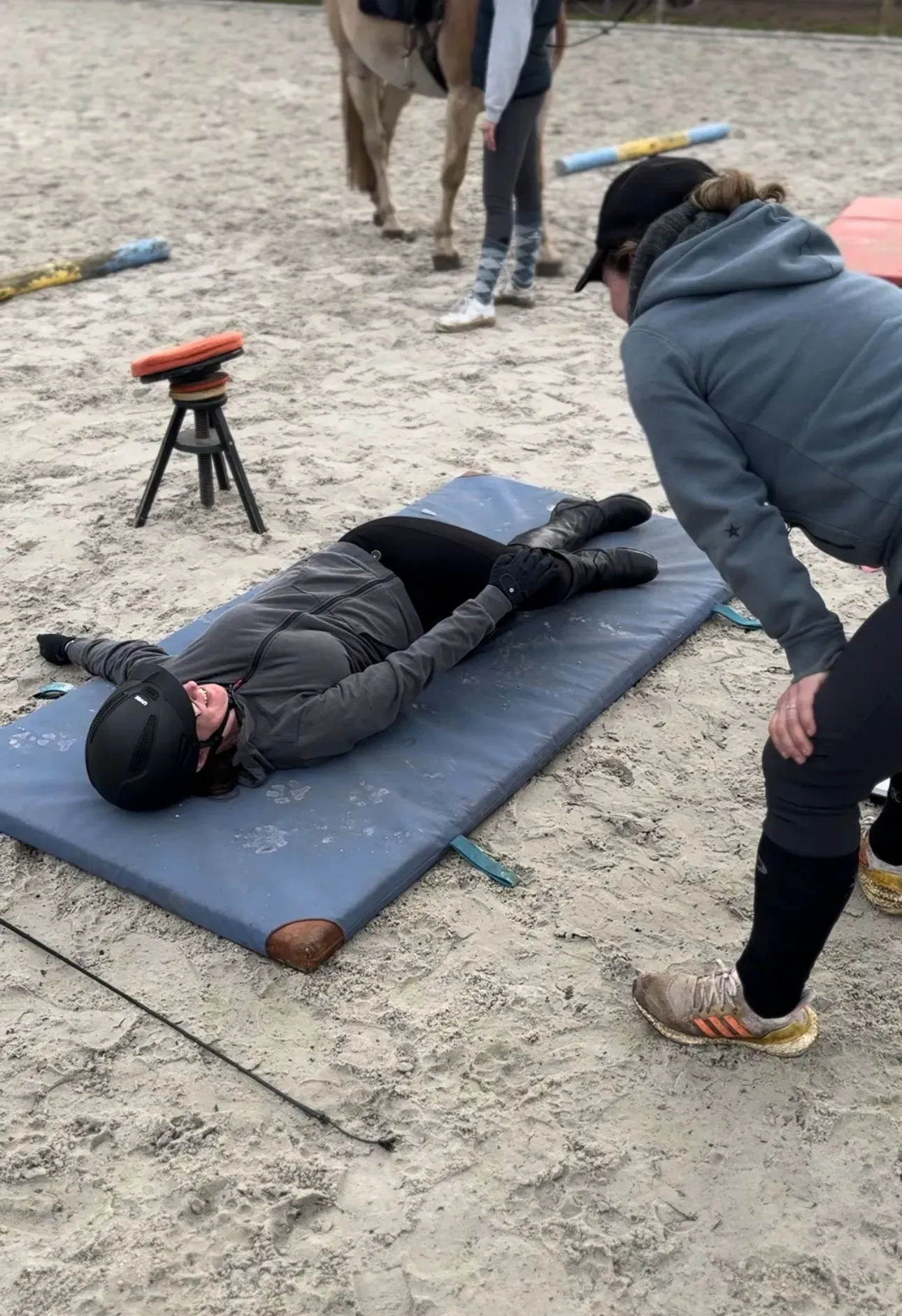 Ein Reiter in Schutzausrüstung liegt flach auf einer Reitmatte, während ein Reitlehrer zusieht; ein Pferd steht in der Nähe.