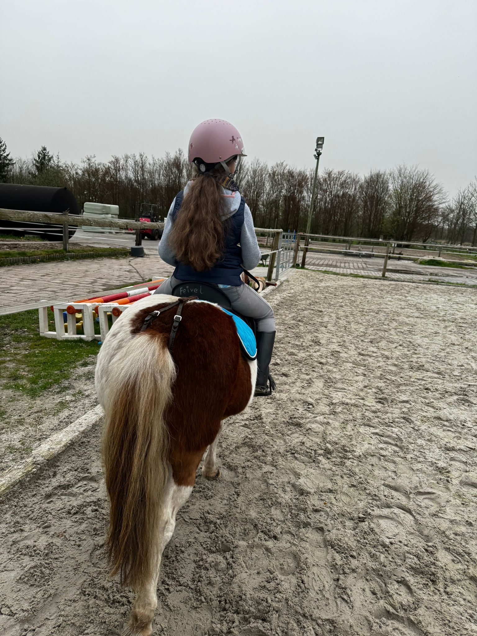 Eine Person mit einem rosa Helm reitet ein braun-weißes Pinto-Pony in einer sandigen Reithalle im Freien.