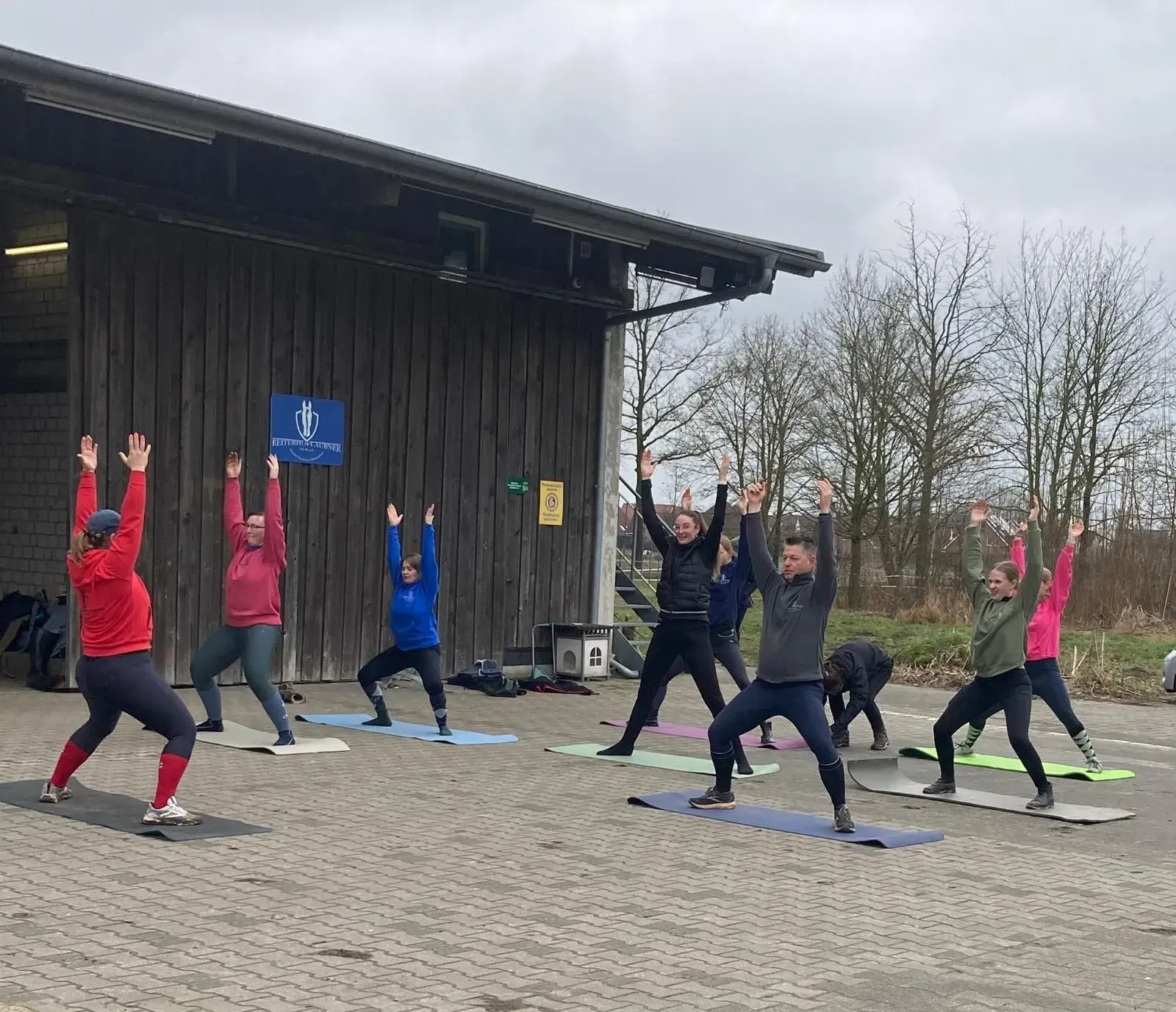 Eine Gruppe von Menschen führt im Freien vor einer Holzscheune Yoga-Übungen auf Matten durch.