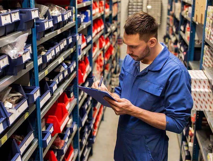 Un trabajador con mono azul revisa el inventario en un portapapeles en un pasillo del almacén.