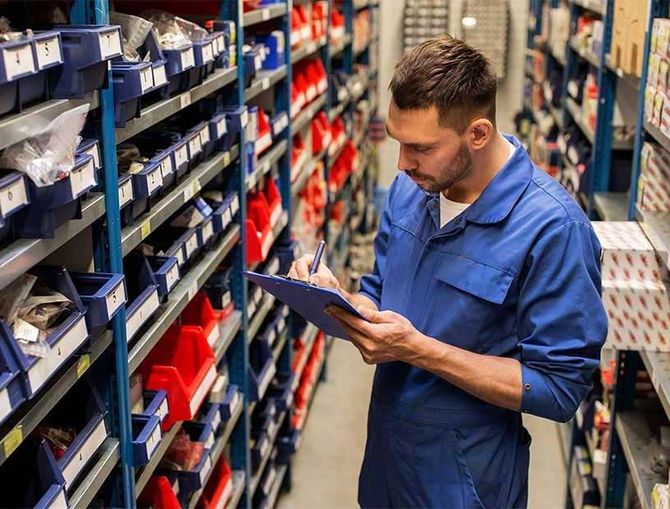 Un trabajador con mono azul revisa el inventario en un portapapeles en un pasillo del almacén.