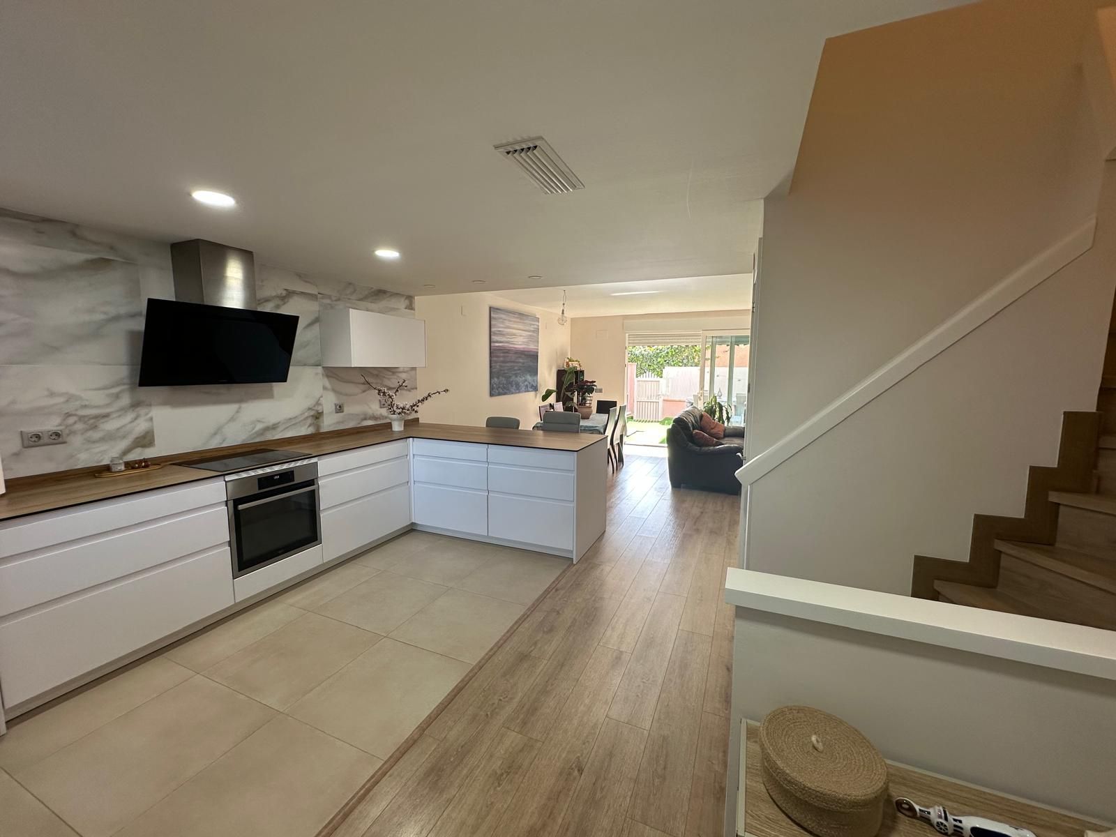 Cocina moderna con gabinetes blancos, encimeras de madera, protector contra salpicaduras de mármol y una sala de estar visible.