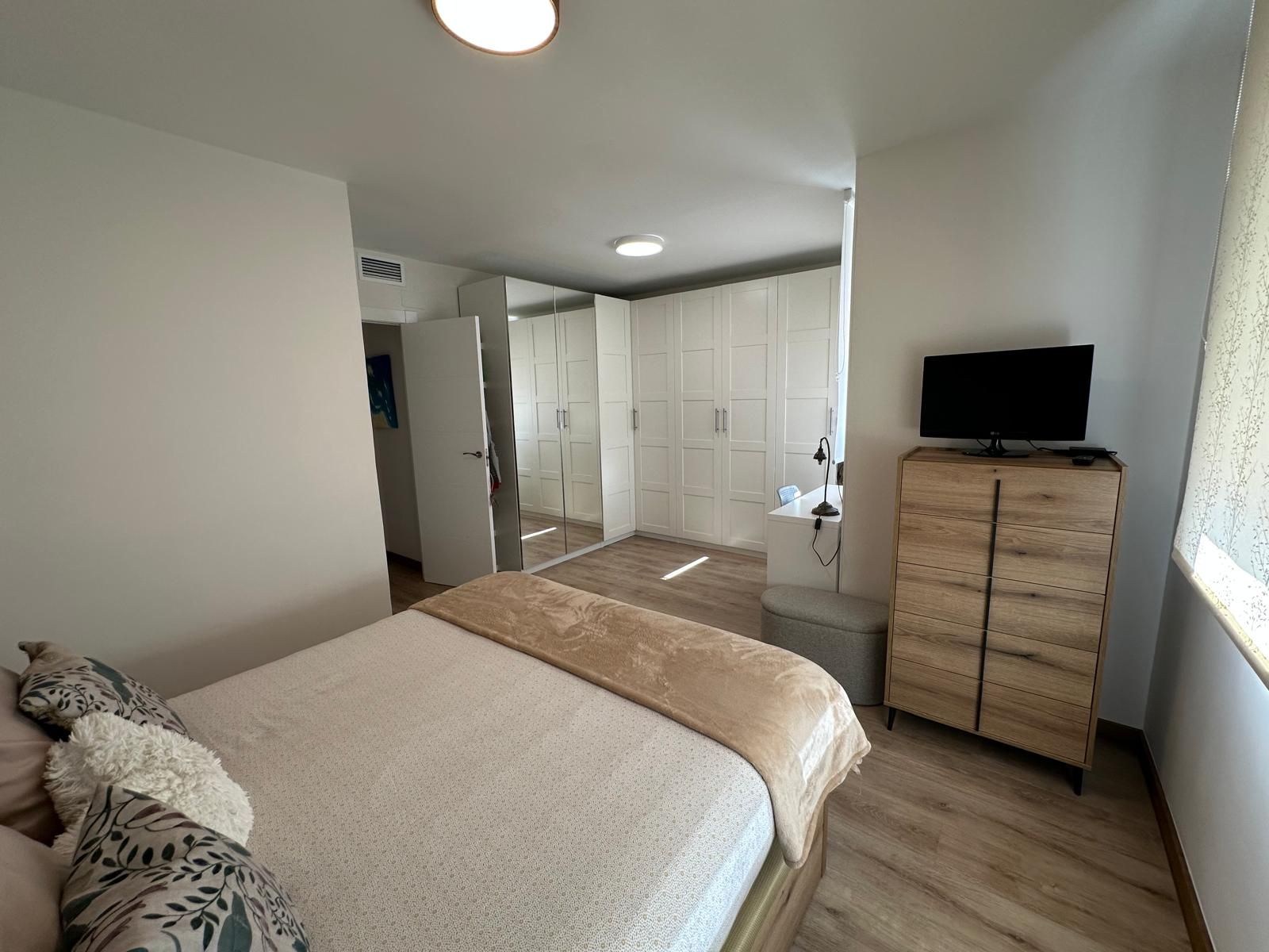 Dormitorio con cama, cómoda, TV y armario blanco con puertas de espejo.