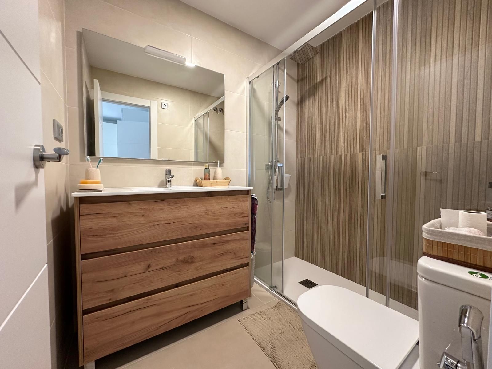Baño con tocador efecto madera, espejo rectangular, ducha de cristal y paredes en tonos neutros.