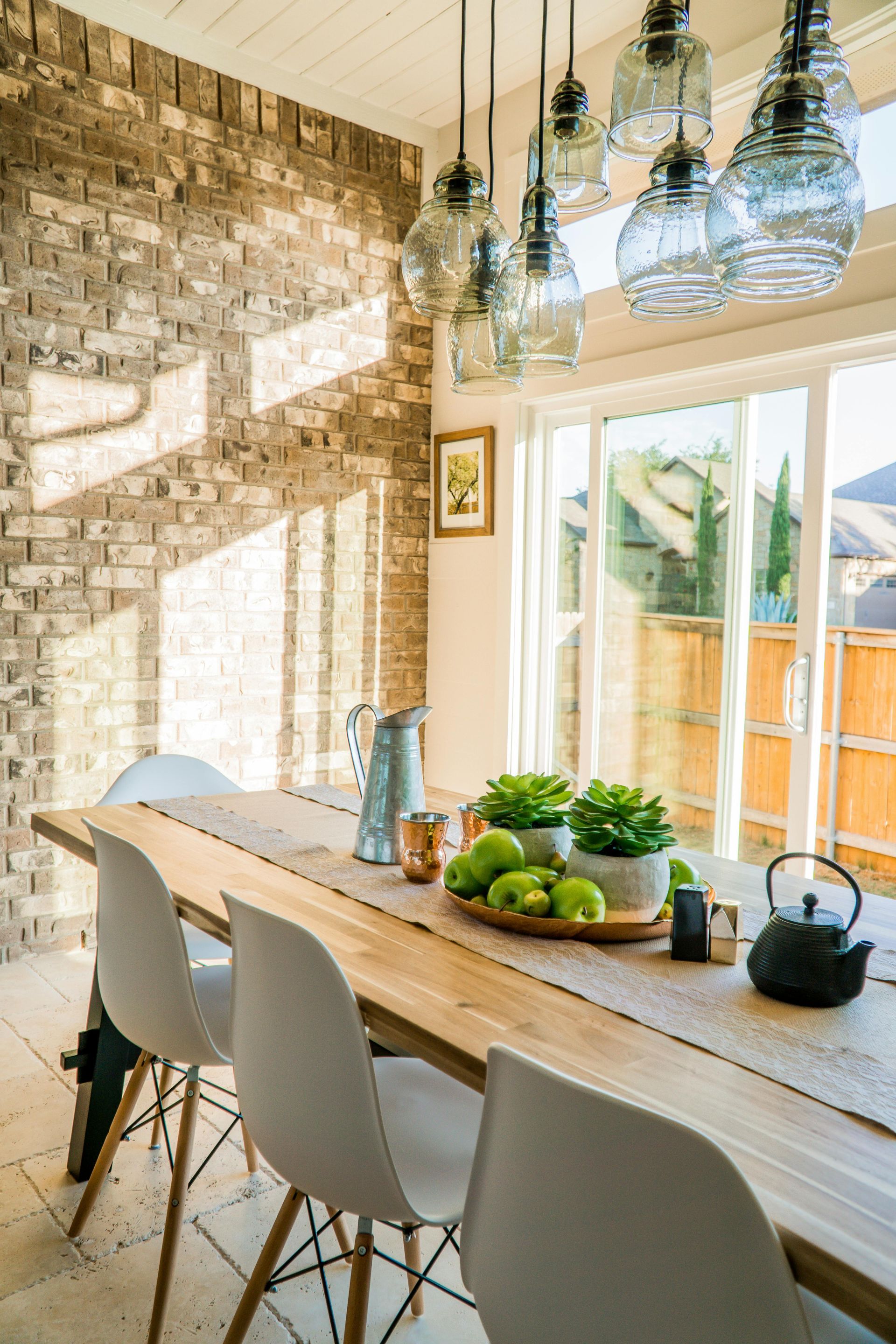 Comedor con mesa larga de madera, sillas blancas, pared de ladrillo y lámparas colgantes de cristal.