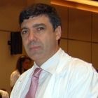 Hombre con bata blanca y corbata rosa; en un espacio interior, mirando hacia el espectador.