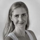 Una mujer con el pelo largo y rubio sonríe, vistiendo una blusa blanca sin mangas, mirando directamente a la cámara.