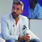 Hombre con blazer azul claro, camisa blanca y vaqueros, sentado, con las manos entrelazadas, mirando fijamente.