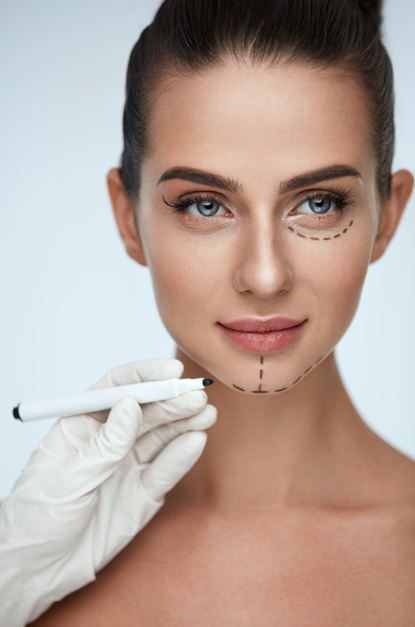 Rostro de mujer con marcas quirúrgicas. Mano enguantada sostiene bolígrafo. Ojos azules, piel clara y fondo neutro.